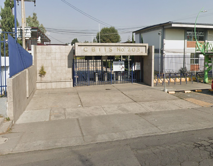 Fotografía en la que se muestra la entrada del plantel, conformada por muros de color gris y una reja azul que da acceso a las instalaciones. Sobre la reja hay un muro que en letras doradas dice Cebetis número 203.